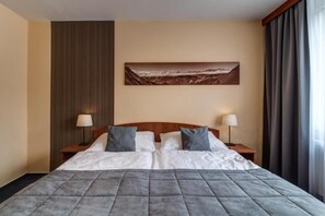 A hotel room with a large bed, two bedside tables with lamps, a painting on the wall, and a window with curtains. - LC Hotel Ostrava (Ostrava)