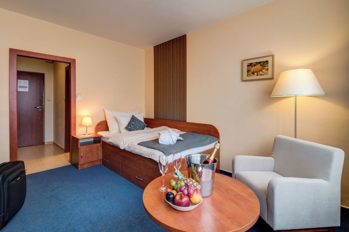A hotel room with a bed, a bedside table with a lamp, a chair, a small table with fruit and champagne, and a door.