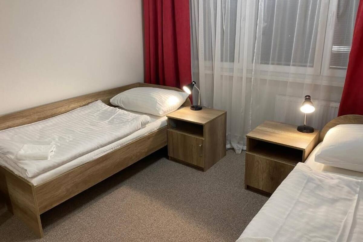 A small, neatly arranged bedroom with two single beds, wooden nightstands, and a window with sheer curtains.