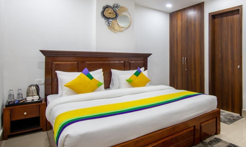 Une chambre avec un lit en bois, une literie blanche et des coussins colorés. On y trouve une table de chevet avec une bouilloire et des verres, ainsi qu'une armoire en bois.