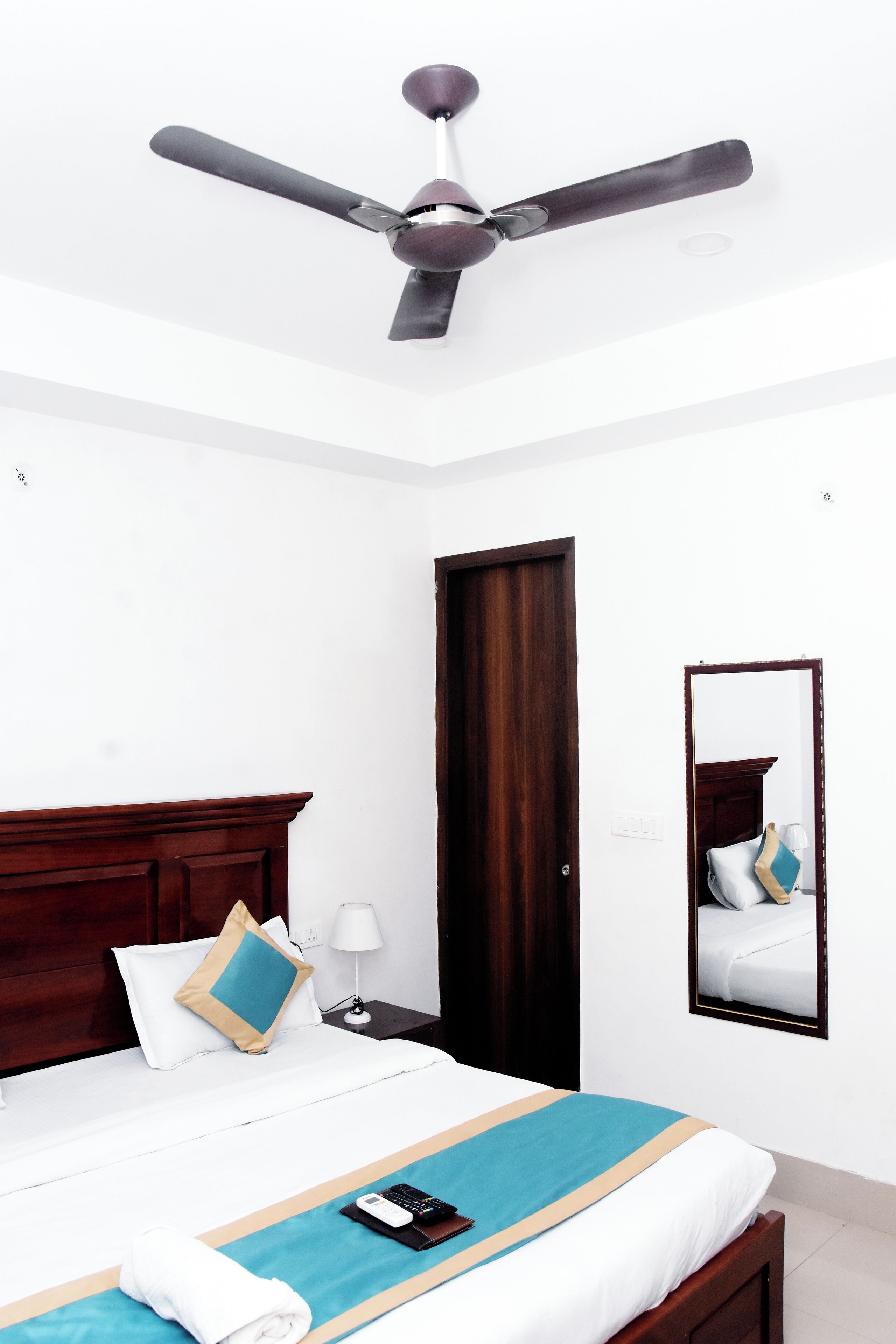 A hotel room with a bed, a ceiling fan, a wooden door, a nightstand, a lamp, a mirror, and a towel on the bed.