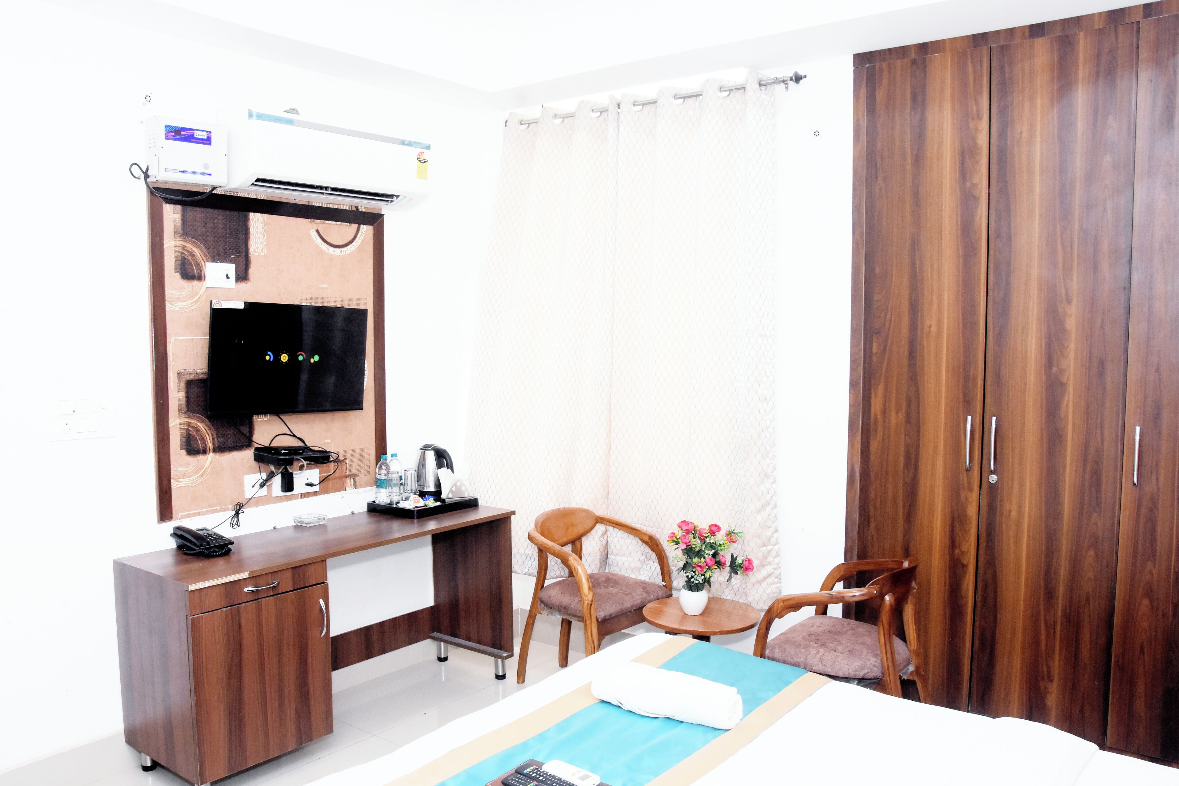 A hotel room with a wooden desk, a bed, a chair, a small table with a vase of flowers, and a wardrobe.