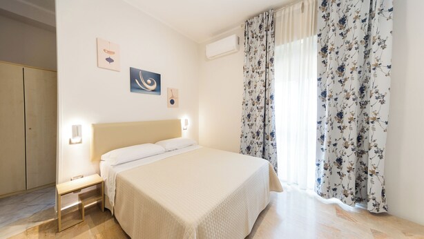 A bedroom with a bed, bedside table, wall art, and a window with floral curtains. - Hotel Dalì near Pompei - Naples (Scafati)