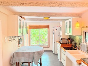 Una cocina con mesa de comedor, armarios, un refrigerador y una ventana con follaje verde afuera.