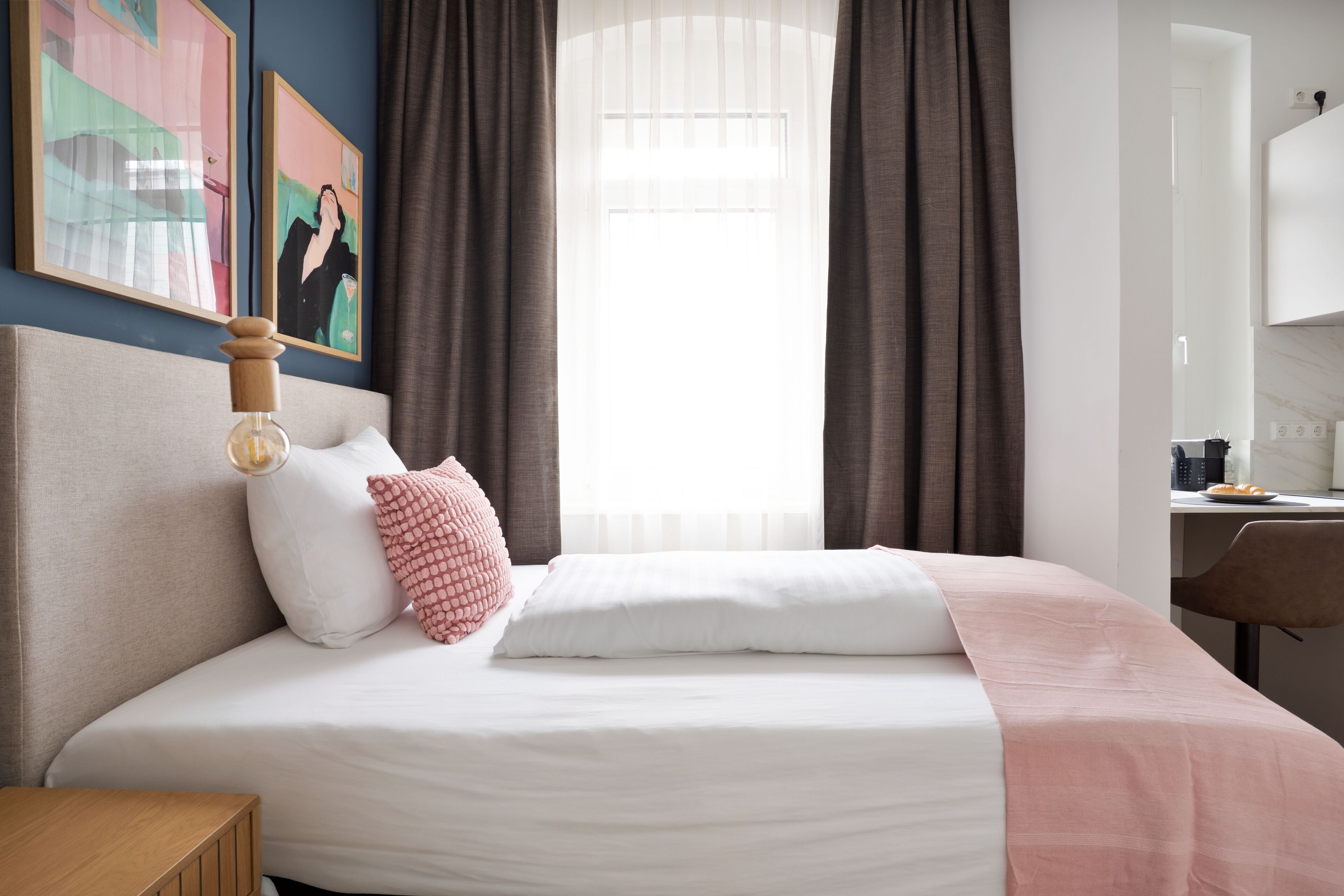A neatly made bed with white and pink bedding, a wooden headboard, and a bedside table with a lamp.