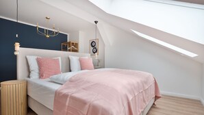 Ein Schlafzimmer mit schrägverdachter Decke, einem Bett mit rosa Decke, einem hölzernen Nachttisch, einer Pendelleuchte und einem Wandregal.