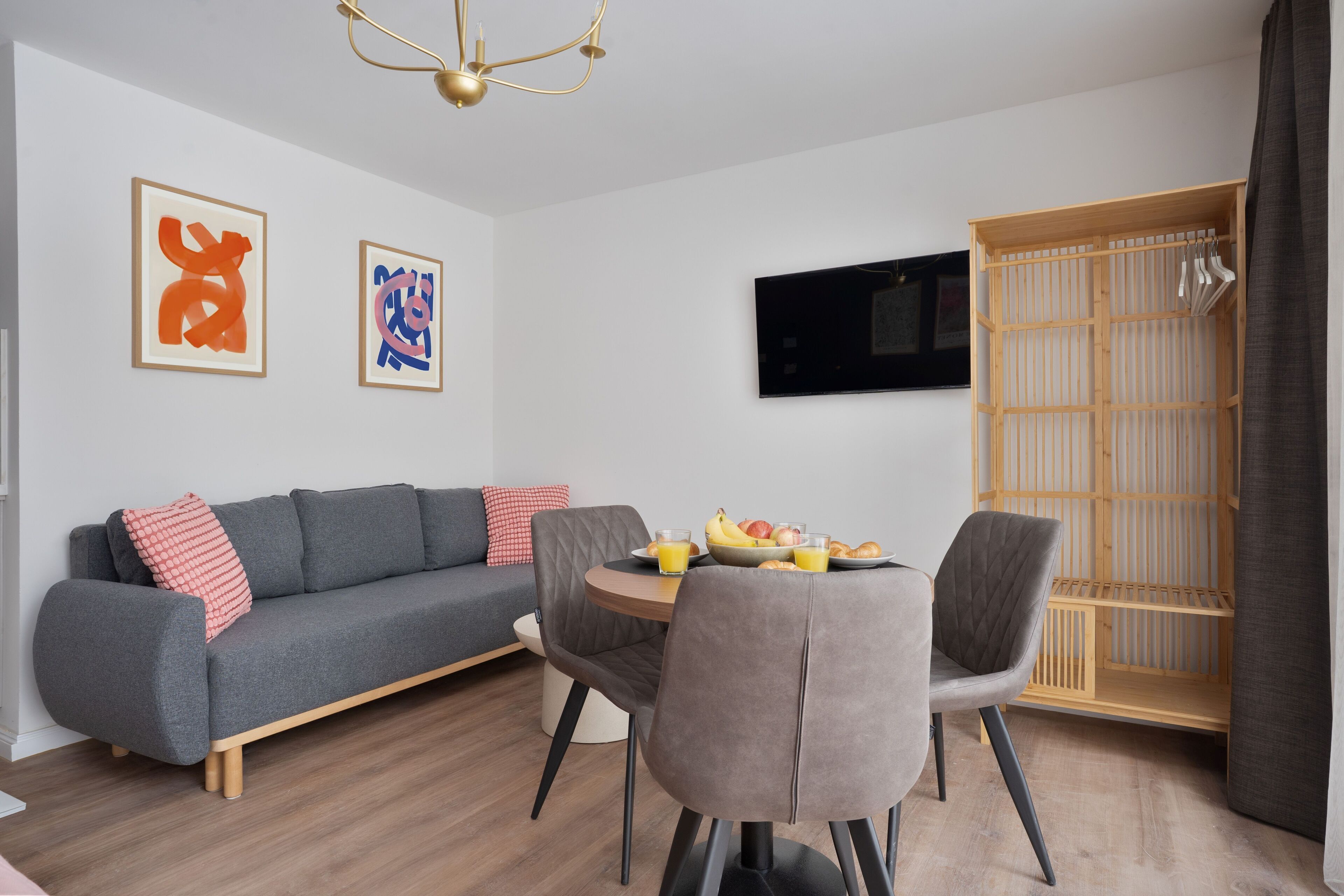 A modern living room with a grey sofa, a dining table with chairs, and a TV mounted on the wall.