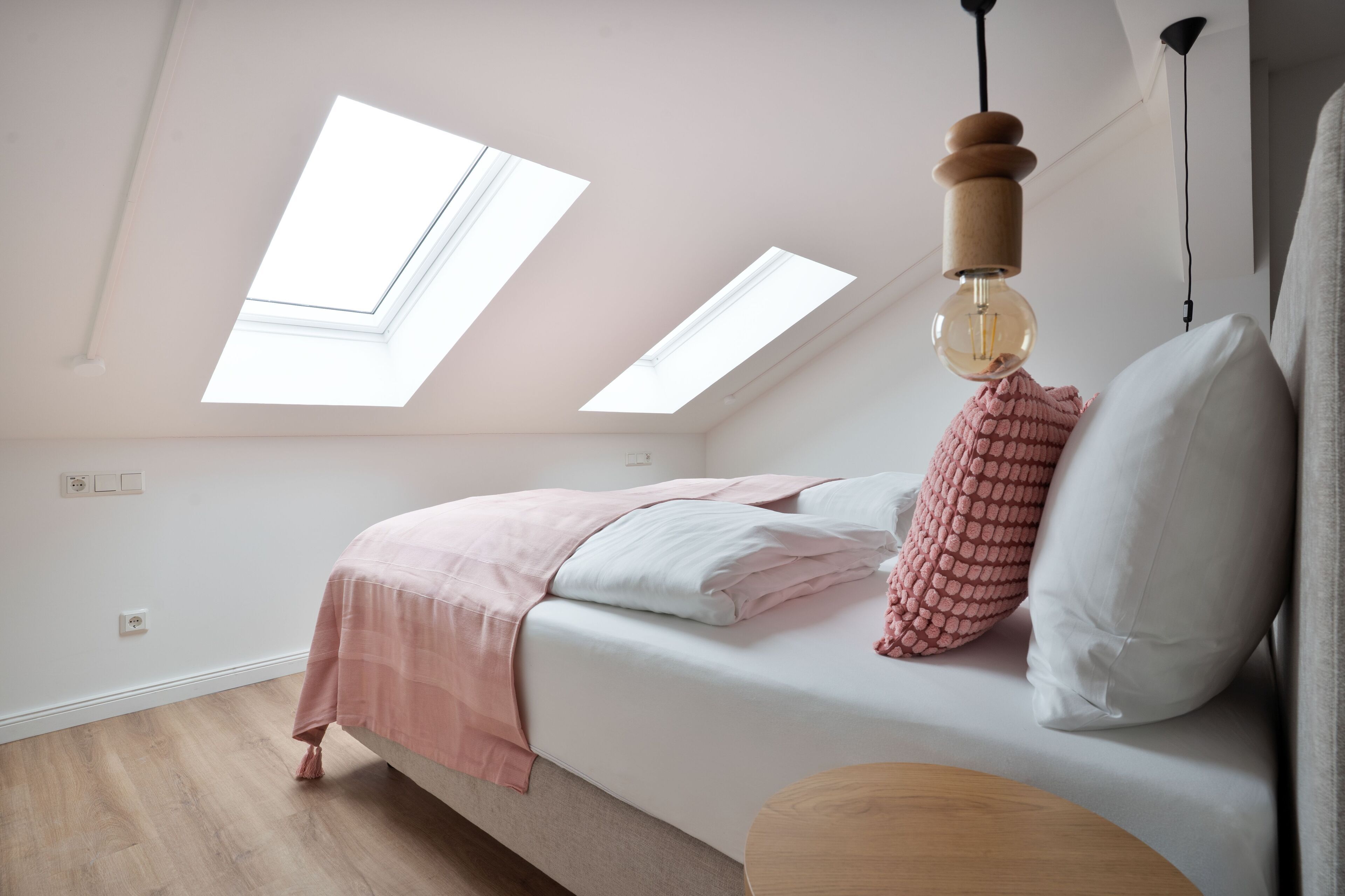 A bedroom with a skylight, a bed with pink and white bedding, and a wooden side table.