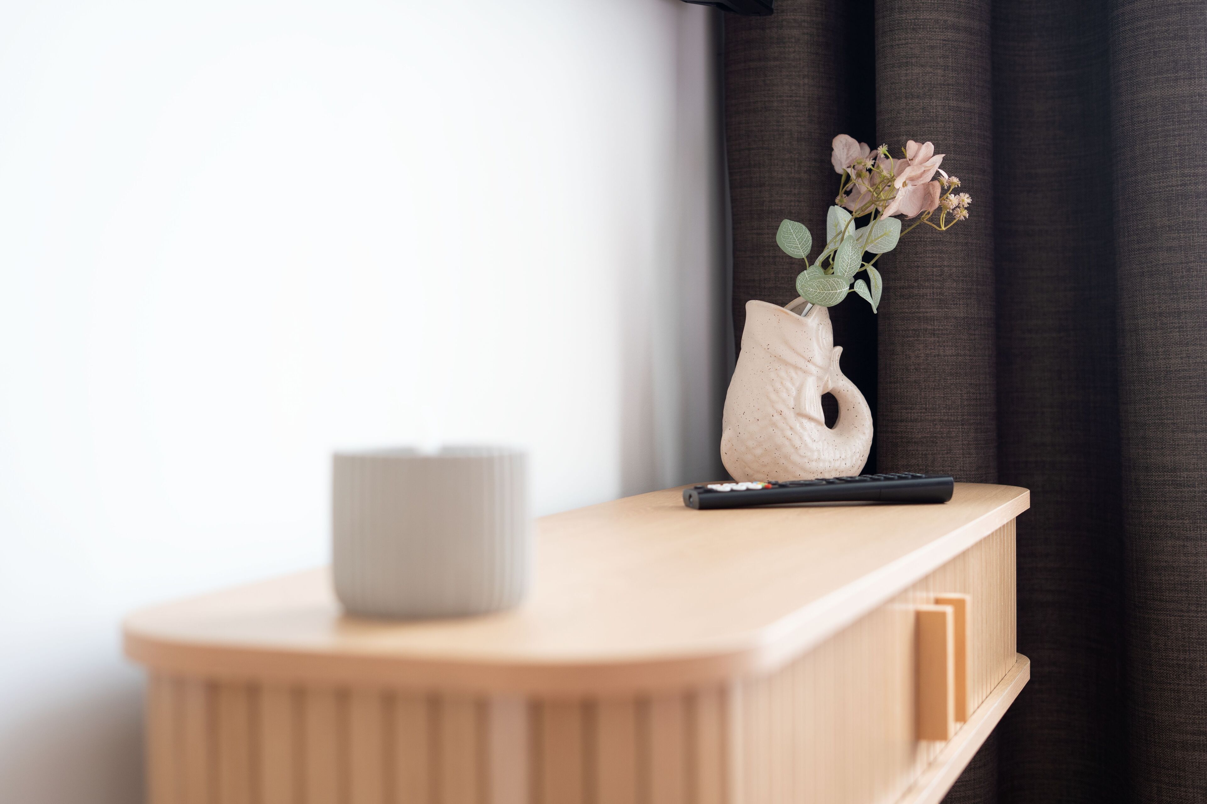 Une table de chevet en bois, ornée d’un vase de fleurs, d’une télécommande et d’une bougie.