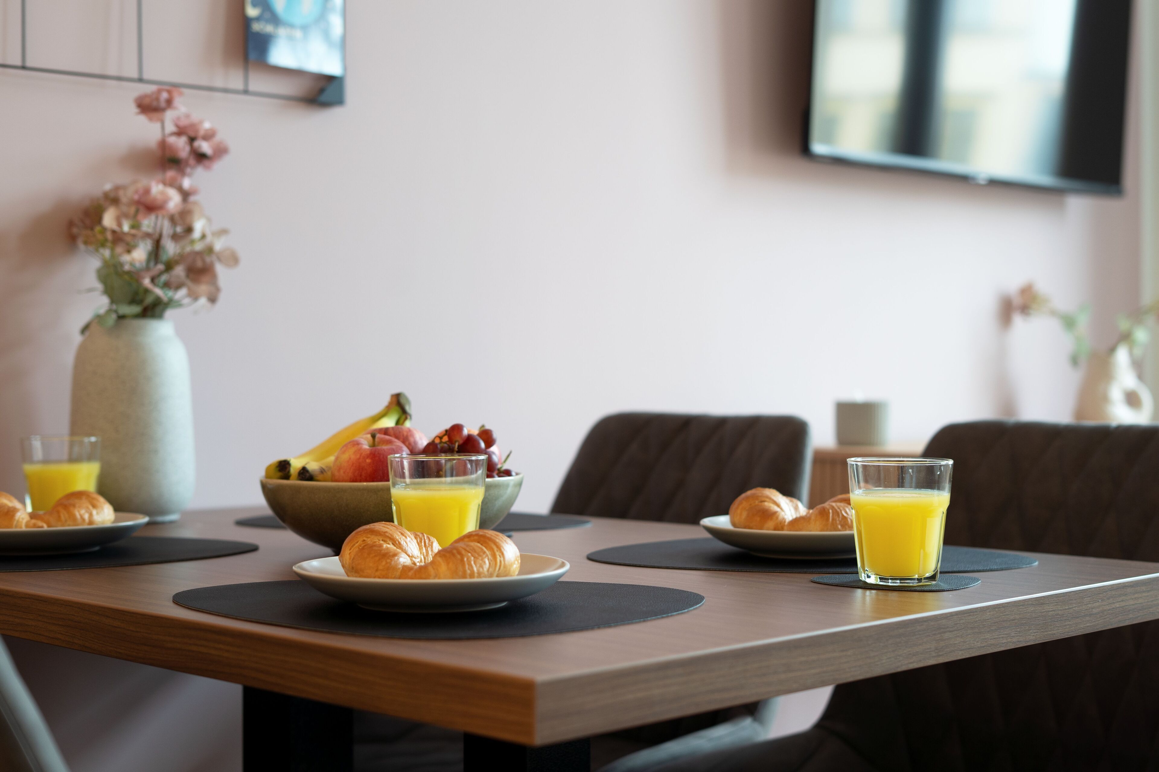 Une table de petit-déjeuner avec des croissants, un panier de fruits et un verre de jus d’orange.