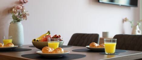 Une table de petit-déjeuner avec des croissants, un panier de fruits et un verre de jus d’orange.