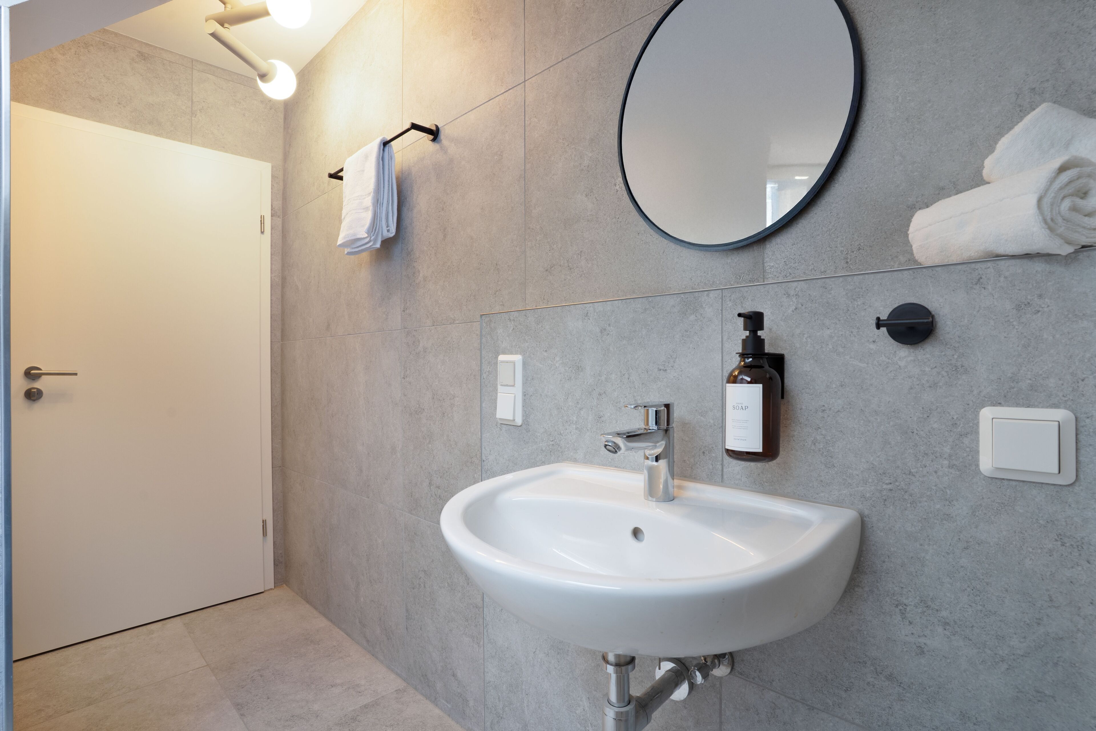 Un baño moderno con un lavamanos blanco, un espejo redondo y un perchero para toallas.