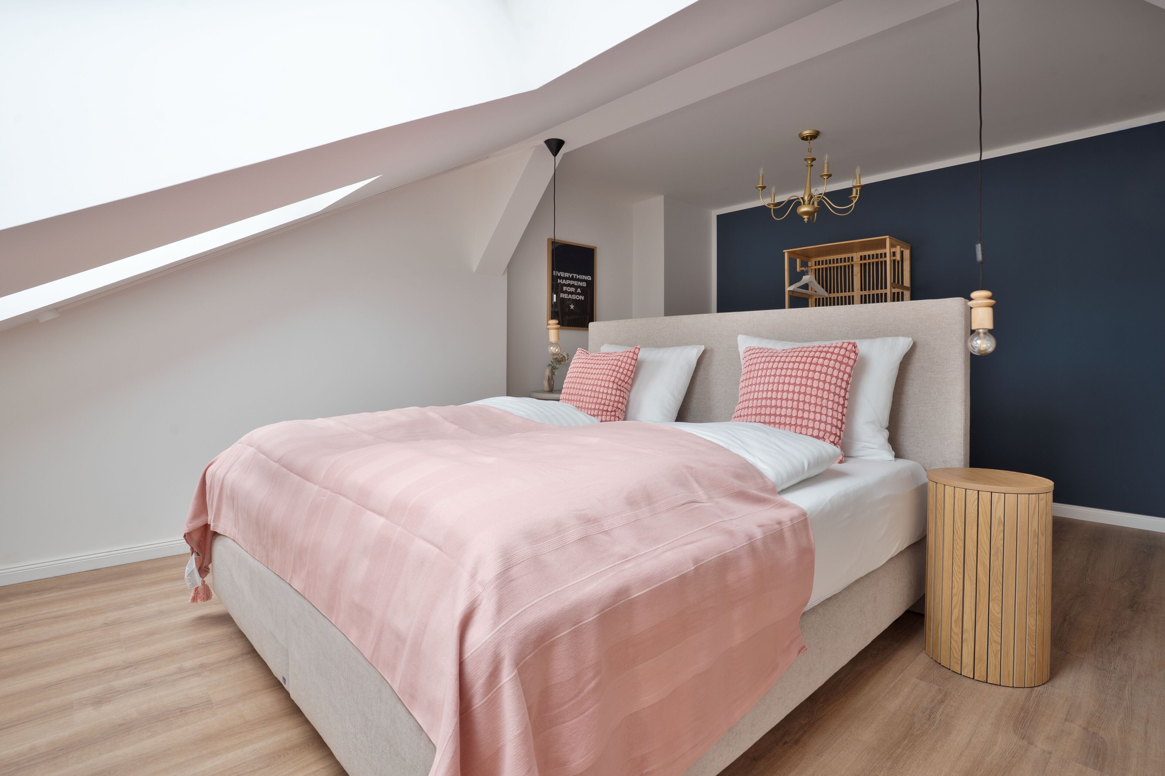 Een slaapkamer met een schuine kap, een bed met roze beddengoed, een houten nachtkastje en een wandlamp.
