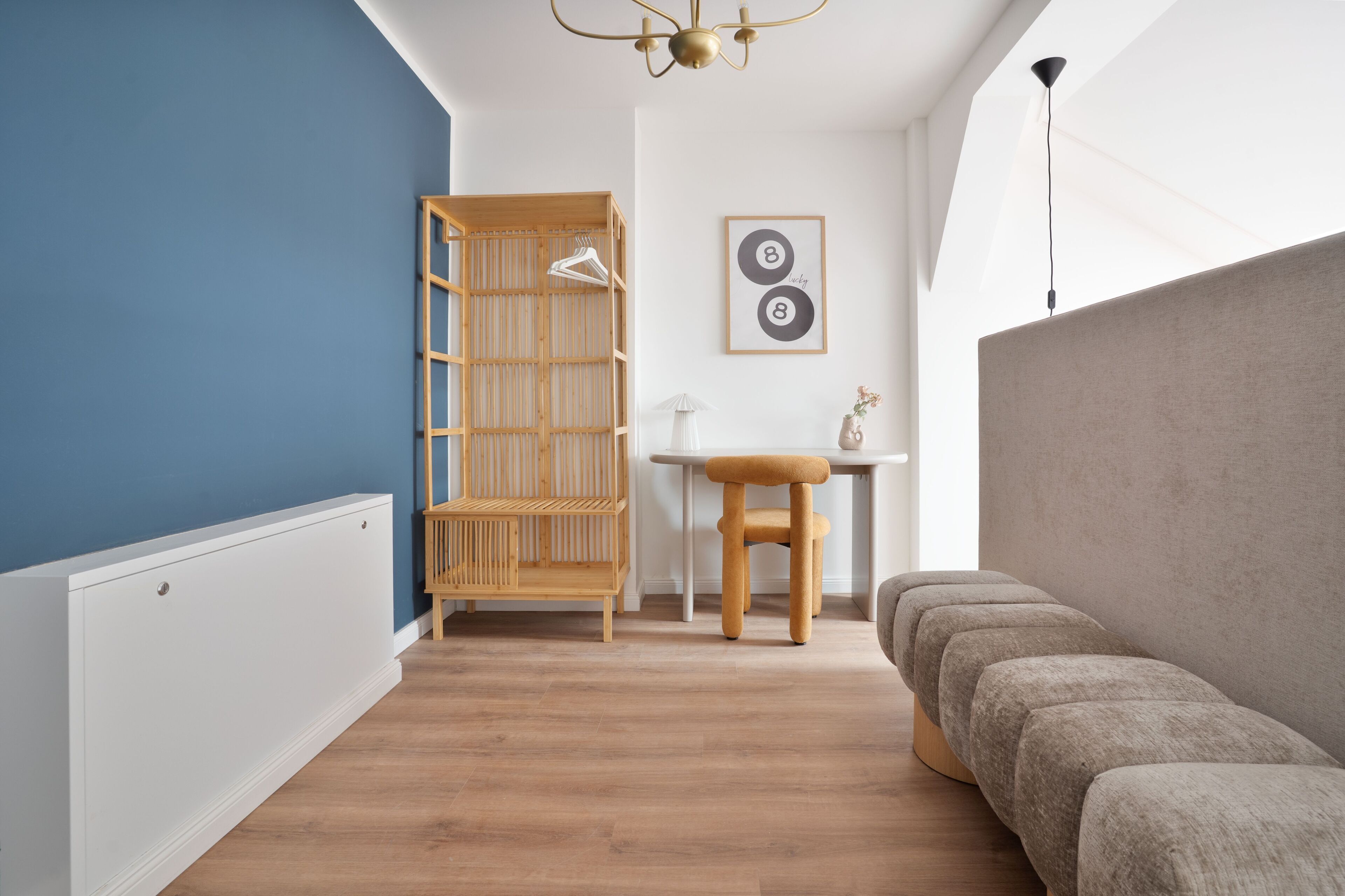 Una habitación moderna con una pared azul de acento, un estante de madera, un radiador blanco, una pequeña mesa con una silla y un sofá gris.