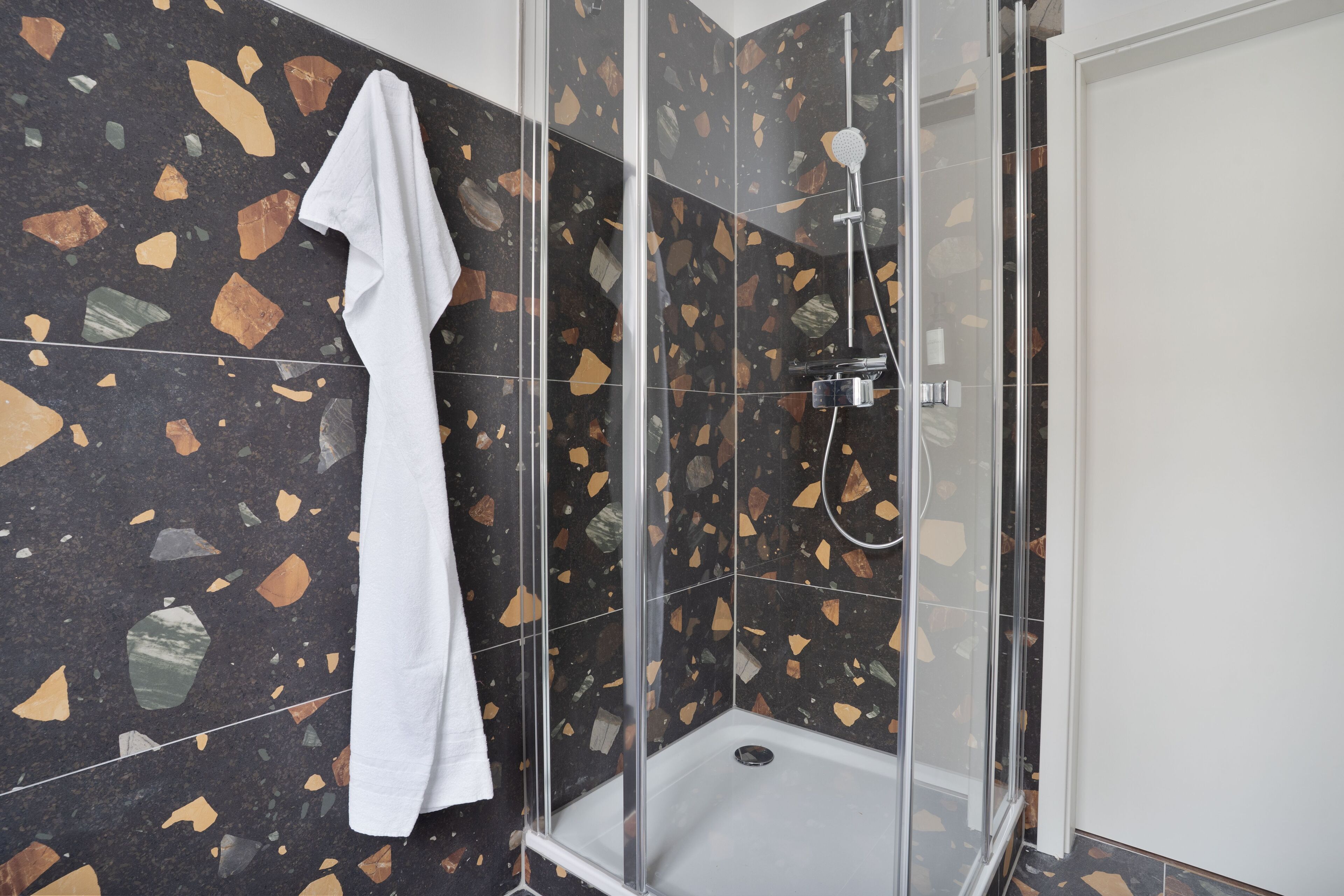 A shower with a glass door, a white towel hanging on the wall, and terrazzo tile flooring.