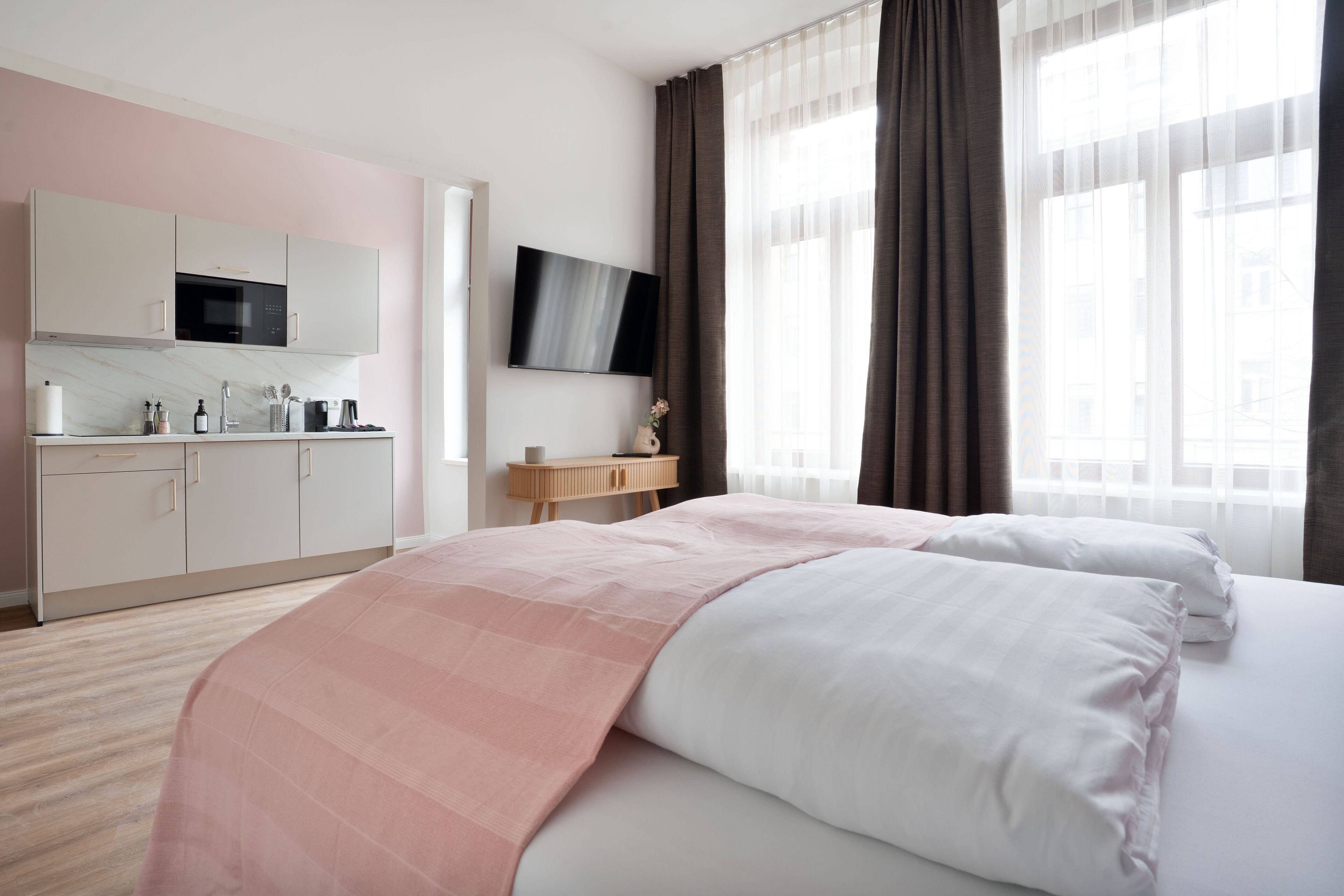 Quarto de hotel com cama, televisão, área de cozinha e janela com cortinas.