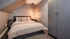 Une chambre avec un plafond en pente, un lit recouvert d’une couverture beige, une table de chevet, une armoire et un luminaire suspendu.