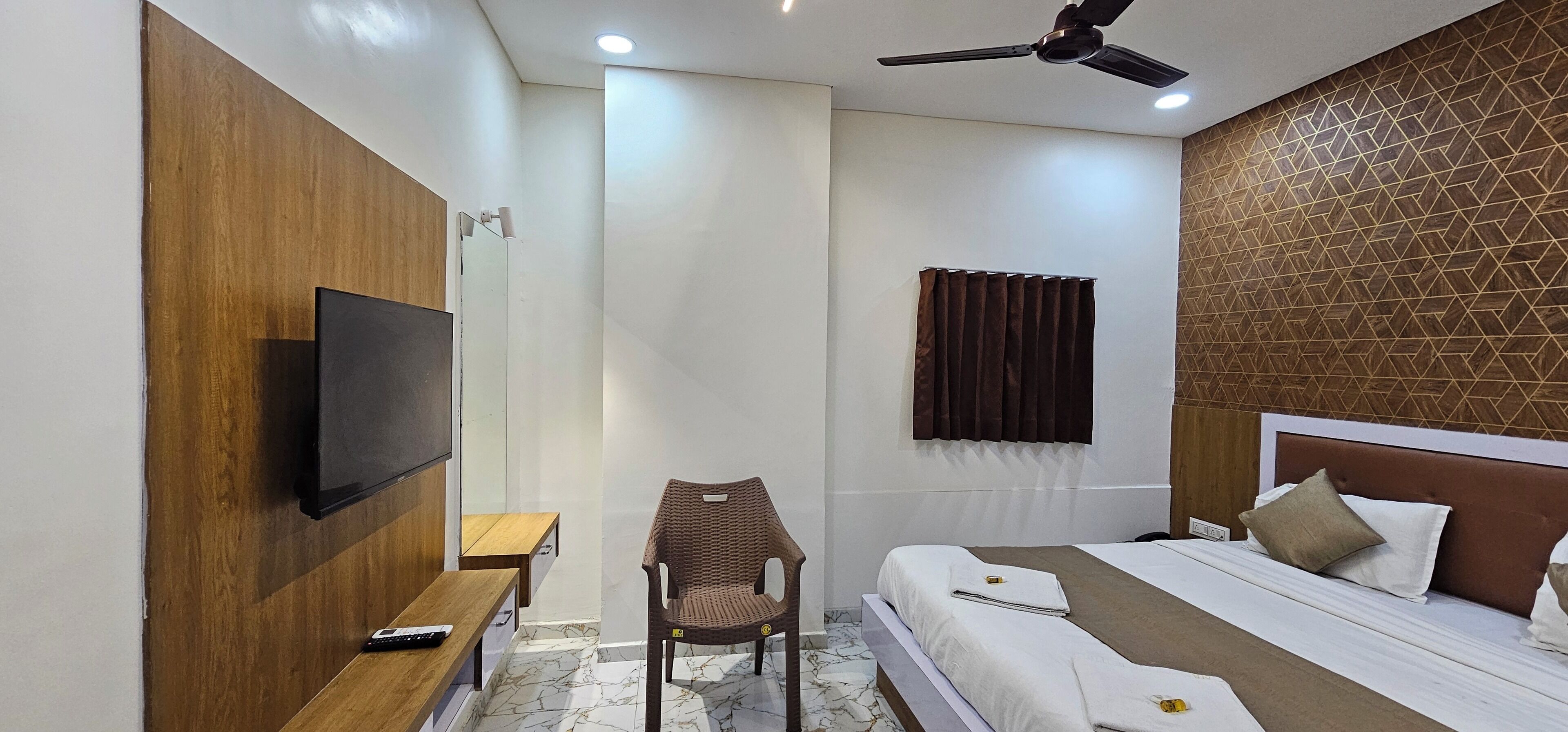 A hotel room with a bed, a TV mounted on the wall, a chair, a small table, and a ceiling fan.