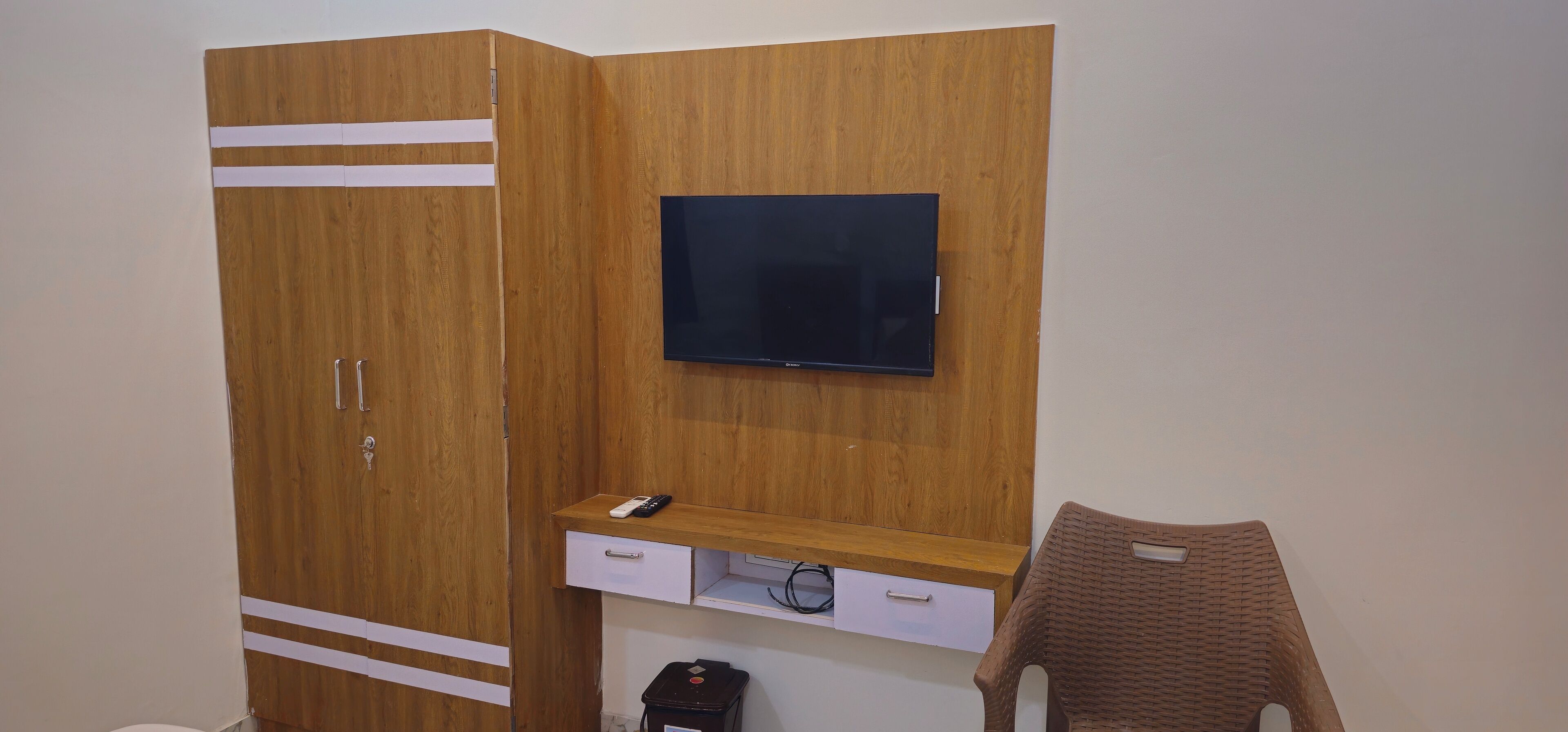 Une pièce comprenant une armoire en bois, un téléviseur fixé au mur, un petit bureau avec un tiroir et une chaise en osier.