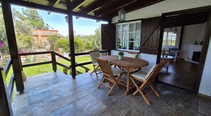 Un porche cubierto con una mesa y sillas de madera, vistas al jardín y una chimenea al fondo.