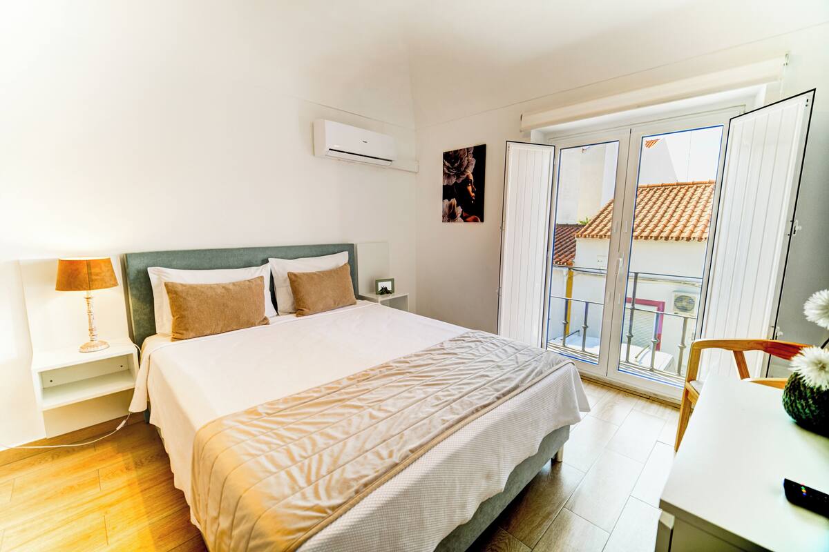 Un dormitorio con cama, mesita de noche, lámpara y vistas al balcón exterior.