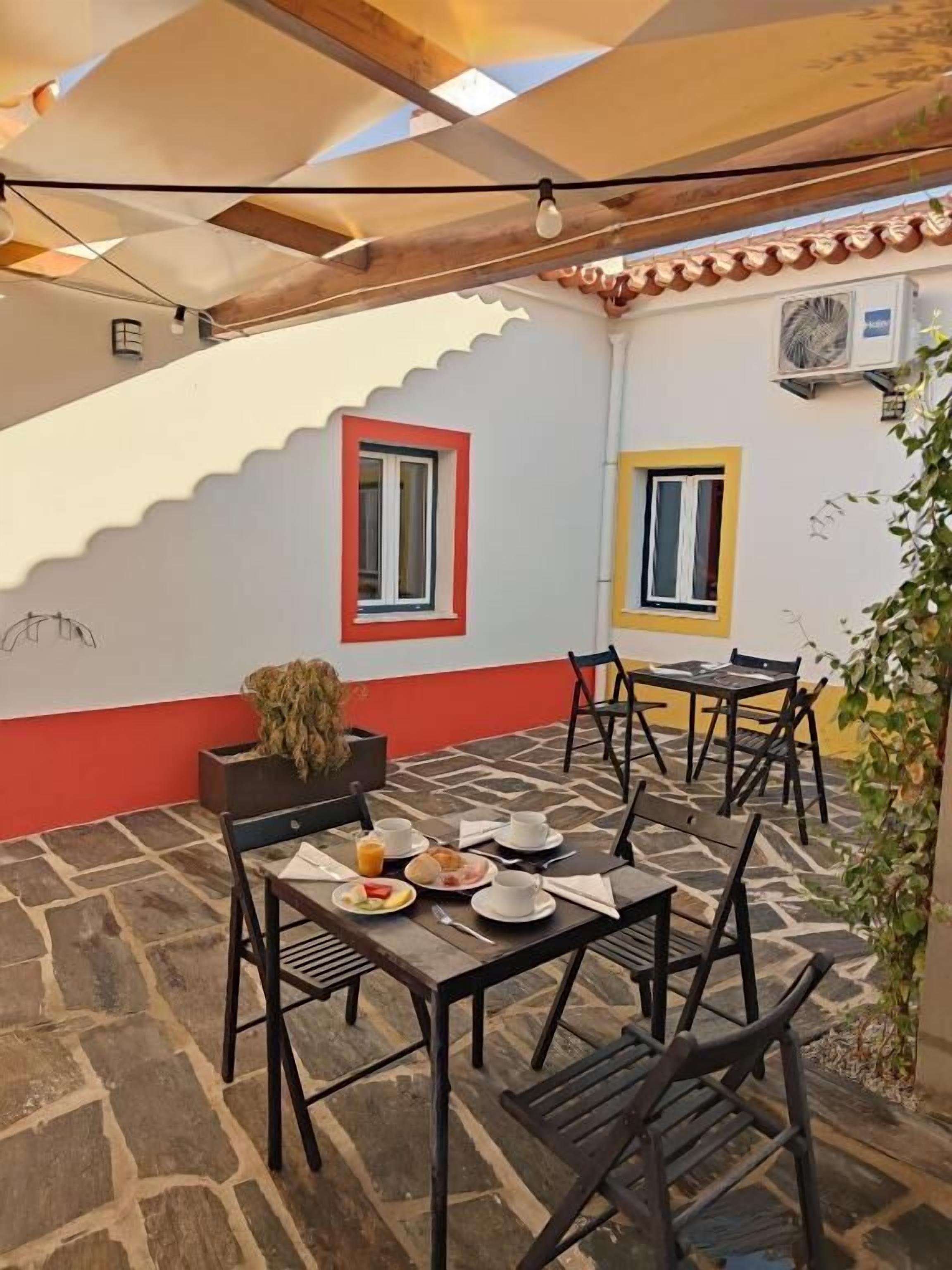 Un espacio de comedor al aire libre con suelo de piedra, mesas y sillas de madera, y dos ventanas con marcos rojos y amarillos.