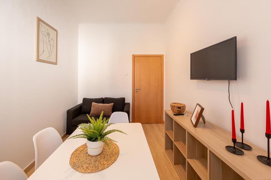 A modern living room with a white sofa, a wooden coffee table with a plant, a TV mounted on the wall, and a wooden shelf with candles.