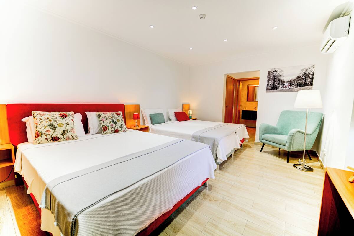 Habitación de hotel con dos camas, cabecera roja, sillón azul turquesa y una lámpara de pie.