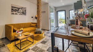 Ein modernes Wohnzimmer mit einem gelben Sofa, einem Holztisch und einem Essbereich mit Tisch und Stühlen. In der Mitte befindet sich ein großer Baumstamm, an der Wand hängt ein Bild und durch eine Glastür ist ein Blick ins Freie möglich.