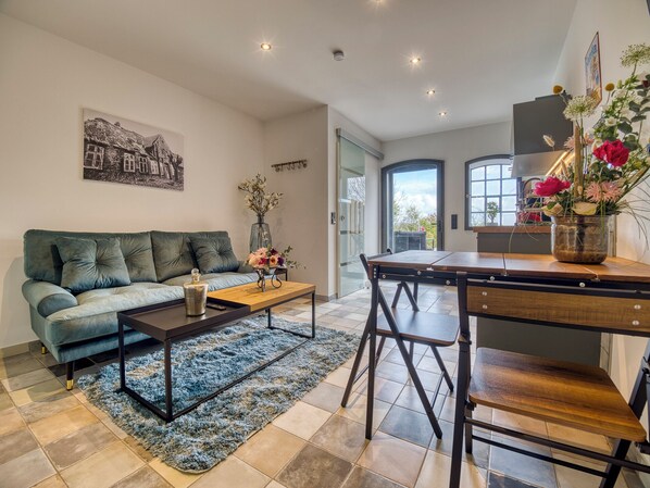 Un salon moderne avec un canapé, une table basse et un coin repas avec des chaises. Il y a une grande fenêtre et une porte donnant sur l’extérieur.