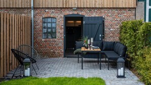 Een houten gebouw met een bakstenen deuropgang, een zwarte deur en een zwarte bank met een tafel en stoelen buiten.