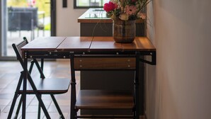 Een eetgedeelte met een houten tafel en stoelen, een bloevas en uitzicht op de omgeving door grote ramen.