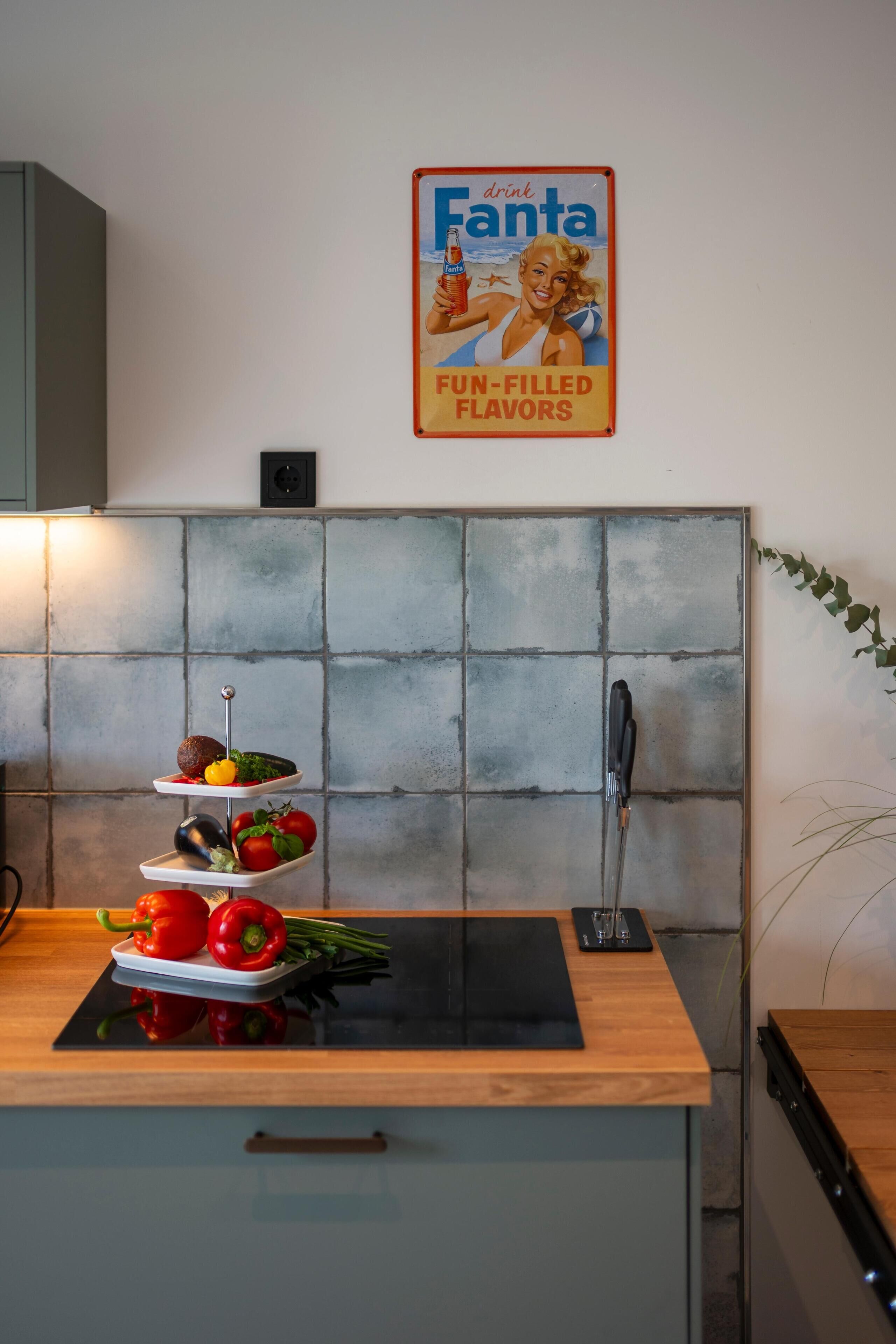 A kitchen with a tiled backsplash, a Fanta poster, and a three-tiered vegetable stand.