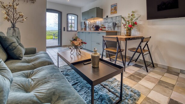 Een moderne woonkamer met een grijze bank, een houten salontafel en een blauw vloerkleed. Door een open deur is een keuken met eethoek zichtbaar.