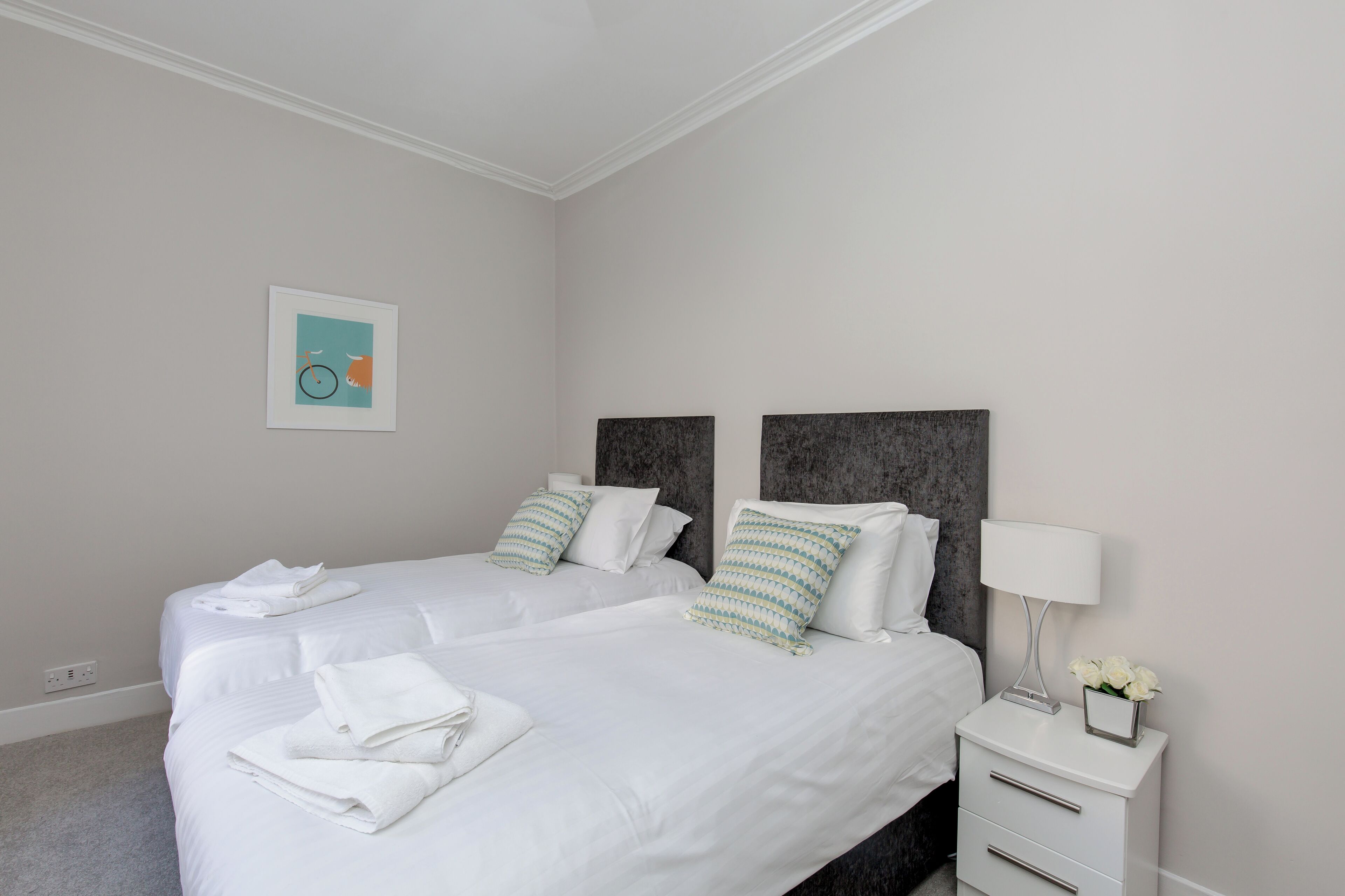 A hotel room with two beds, white bedding, a nightstand with a lamp, and a framed picture on the wall.