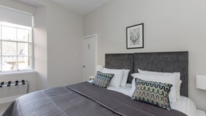 A bedroom with a large bed, two patterned pillows, a framed picture on the wall, and a window with blinds. - Grassmarket Modern Two Bedroom Flat  (Edinburgh)
