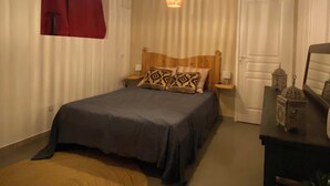 A bedroom with a bed, two hanging lamps, a wooden headboard, a red curtain, a white door, and a small table with decorative items. - The plaid zebra (Entre-Deux)