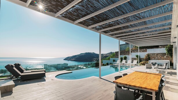 Un área de piscina exterior moderna con sillones de descanso, una mesa de madera y vistas al mar.