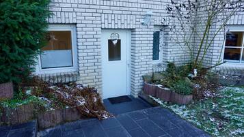 Ein weißes Backsteingebäude mit einer weißen Tür, einem herzförmigen Fenster und einem kleinen Gartenbereich mit Pflanzen und einer Ziegelumrandung.
