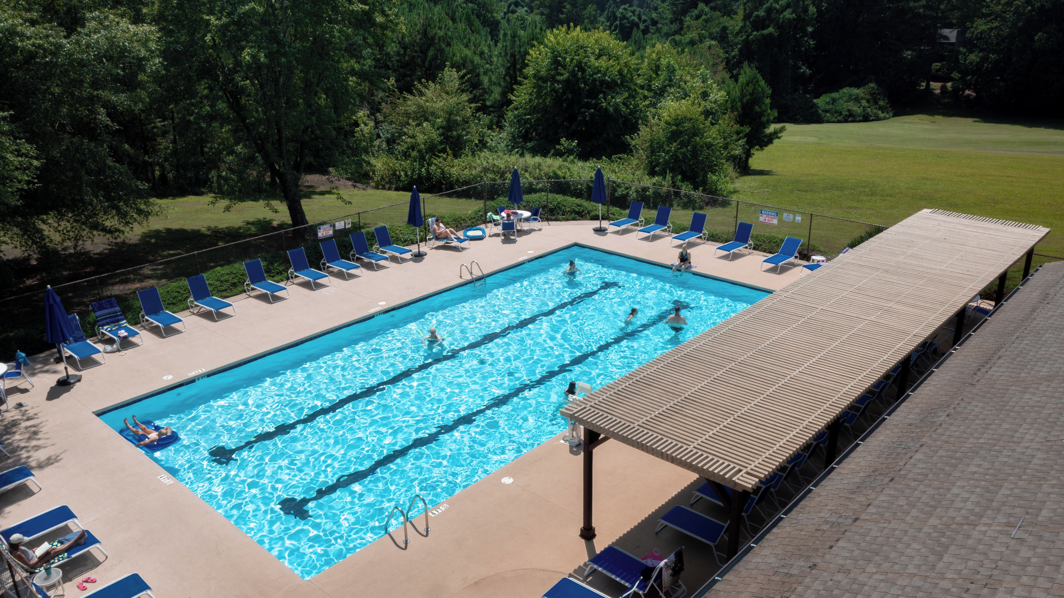 A rectangular swimming pool with multiple lanes, surrounded by lounge chairs and umbrellas, with a covered seating area and a grassy area in the background.