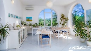 A bright, spacious dining area with a white table set for four, surrounded by potted plants and large windows offering a view of greenery. - Casa Giada nel cuore del Salento (Gallipoli)