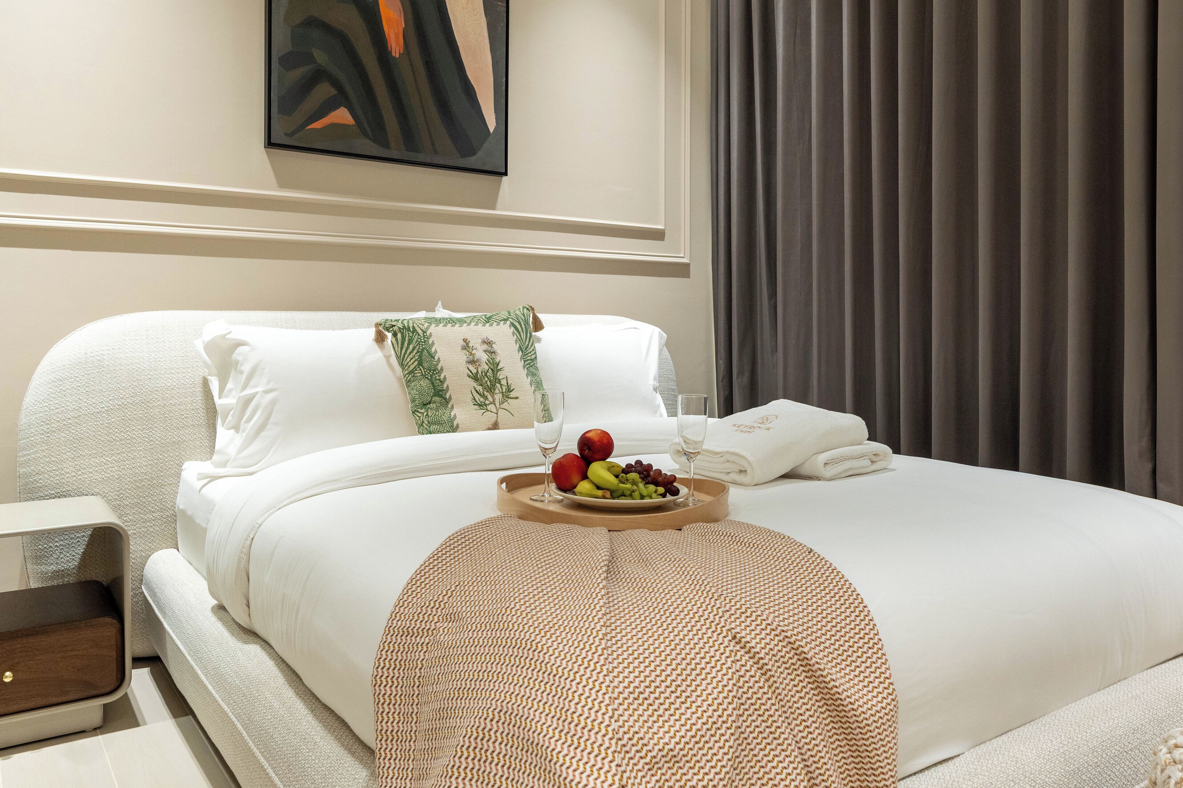 Ein ordentlich gemachtes Bett mit weißer Bettwäsche, eine Obstschale auf dem Bett und ein Nachttisch mit einer Lampe.