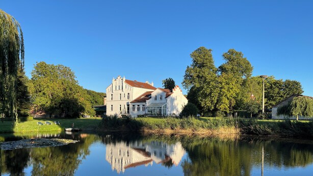Un edificio grande y blanco con techo rojo, rodeado de exuberante vegetación y un lago tranquilo que refleja su imagen.
