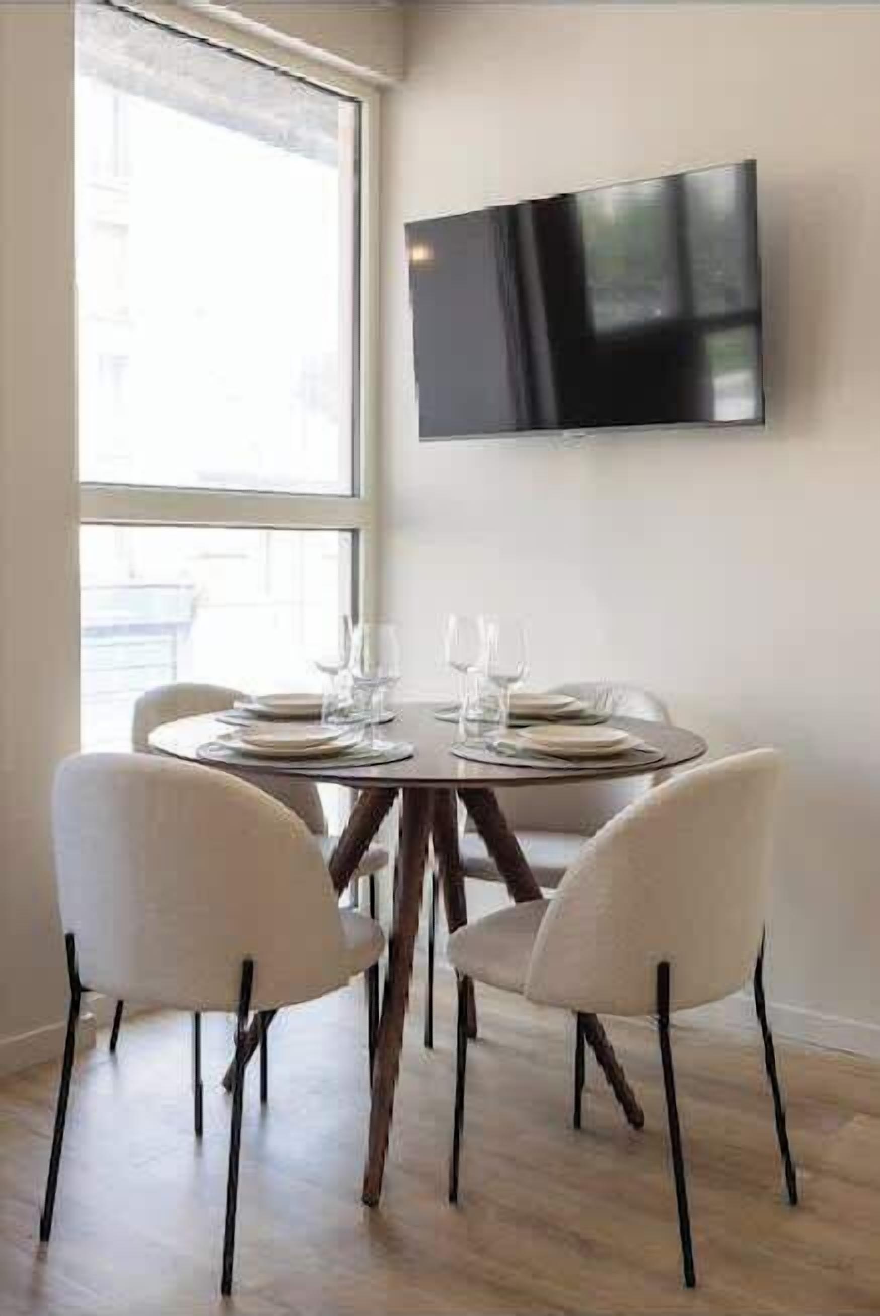 Un coin repas avec une table ronde prévue pour quatre personnes, un téléviseur à écran plat fixé au mur et une grande fenêtre laissant entrer la lumière naturelle.