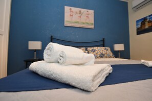 Ein ordentlich bezogenes Bett mit blauer Tagesdecke und einem weißen Handtuch darauf. Zwei Nachttische mit Lampen, eine blaue Wand und ein Gemälde über dem Bett.