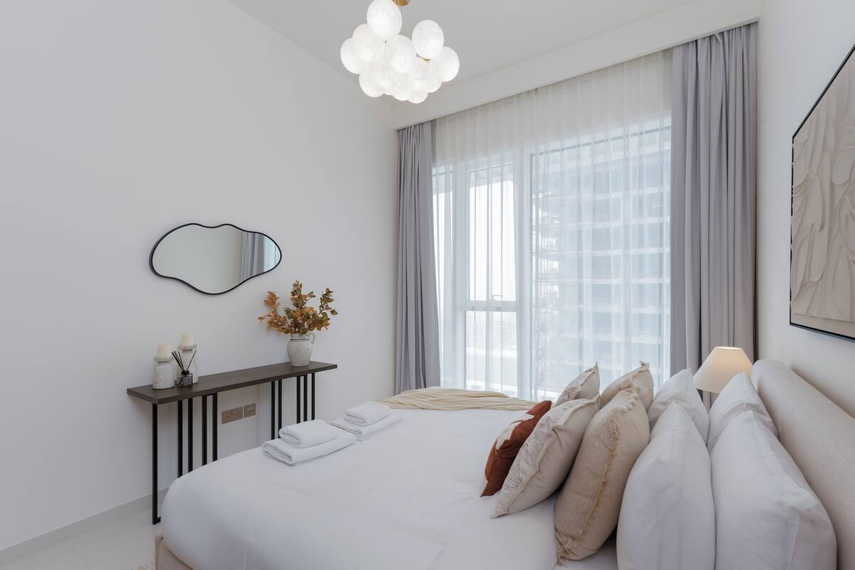 A modern bedroom with a large bed, a bedside table with a vase, and a large window with curtains.