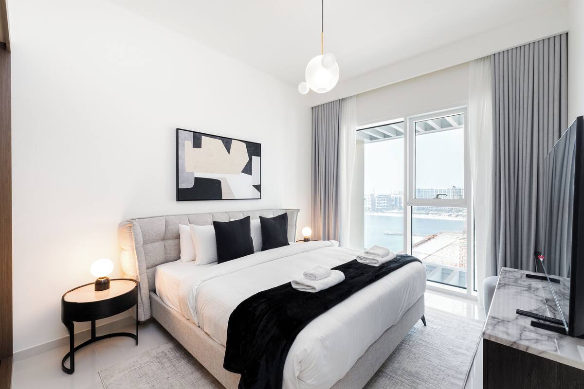 A modern hotel room with a large bed, a television, and a view of the city through large windows.