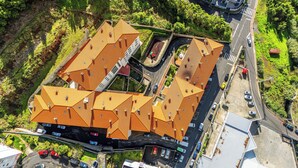 Vue aérienne d’un bâtiment dont le toit présente une structure géométrique particulière, entouré de verdure et bordé par une route où sont garées des voitures.