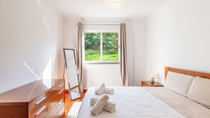 Une chambre à coucher avec un lit, une commode en bois, une table de chevet avec une lampe et une fenêtre avec des rideaux.