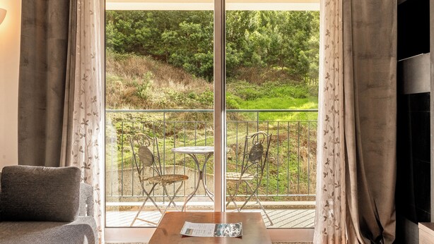 Un salon avec un canapé, une table avec une télécommande et un téléviseur. Il y a un balcon avec une table et des chaises, et une vue sur la verdure à l’extérieur.